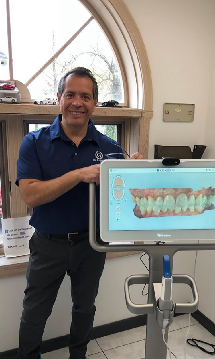 The image shows a man standing next to a dental chair with a digital display screen, holding a model of teeth, smiling and posing for the photo.