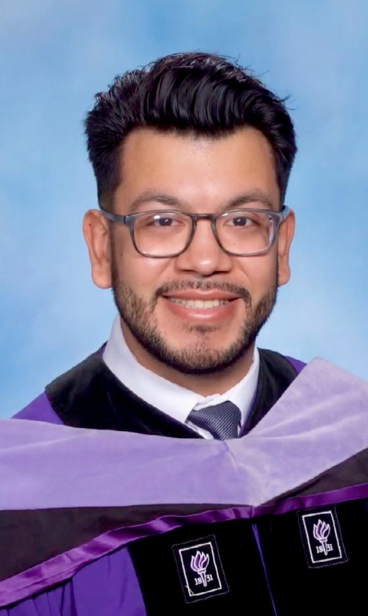 The image shows a man wearing a graduation cap and gown with a purple sash, smiling at the camera.