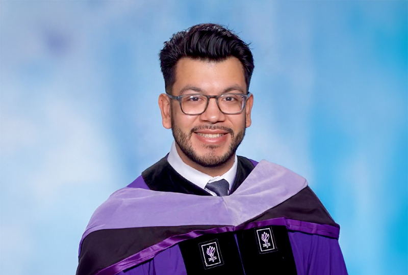 A man wearing glasses, smiling, and dressed in a graduation gown with a purple sash, indicating academic achievement.
