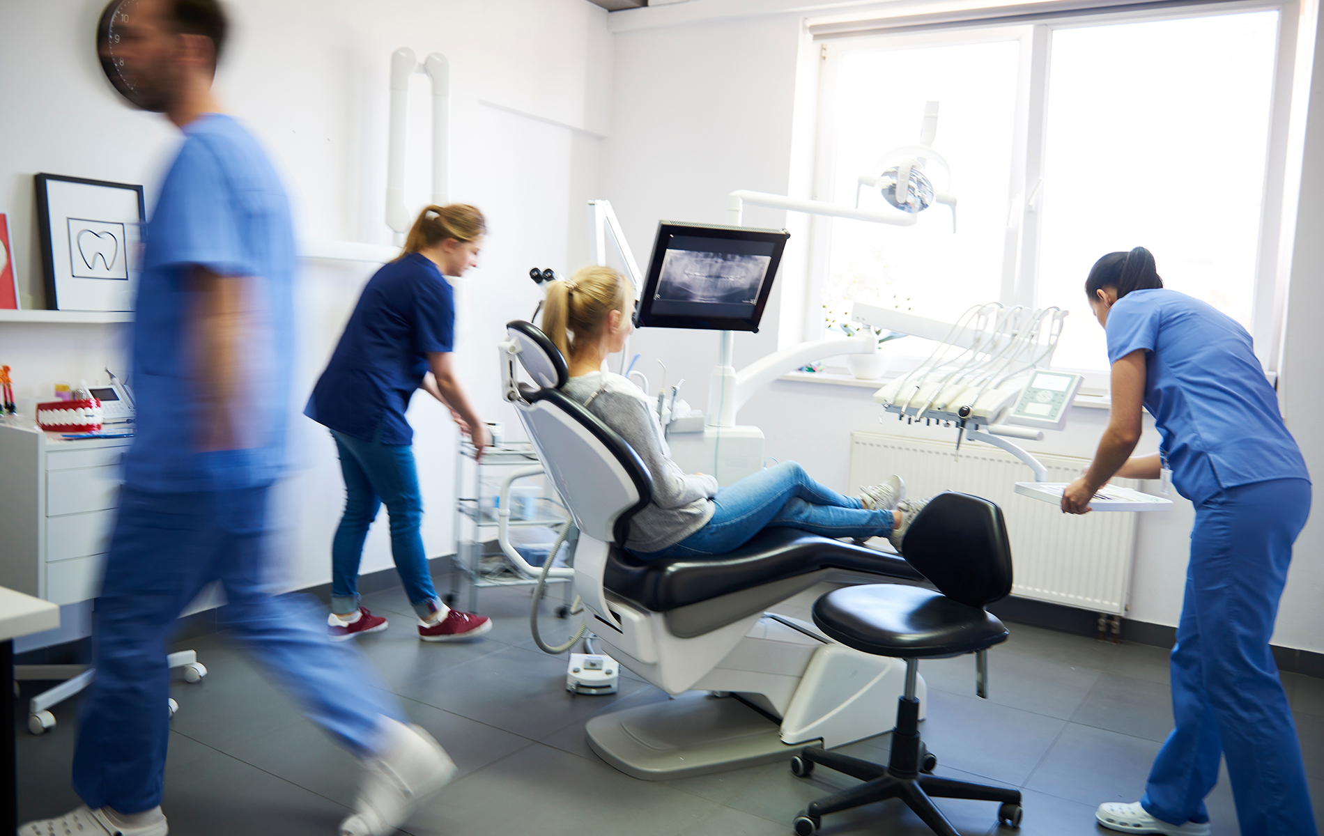 The image shows three photos of healthcare professionals in a dental office setting, with one individual walking past a dental chair and another standing next to an examination table, both dressed in medical scrubs, while the third photo depicts a dental professional sitting at a desk with a computer monitor.