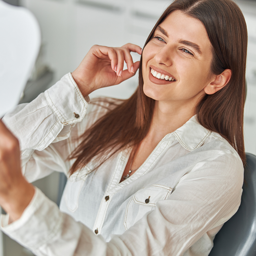 A smiling woman with long hair seated at a desk, holding a phone to her ear, looking towards the camera.