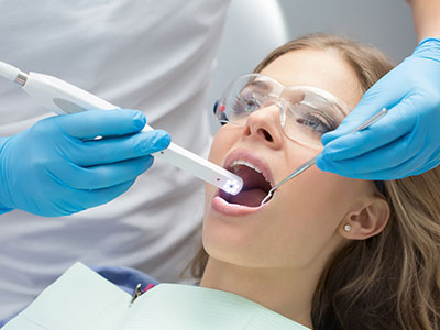 The image shows a woman receiving dental care  she s seated with her mouth open wide, and a dental professional is using an instrument to examine her teeth while wearing protective gloves and goggles.